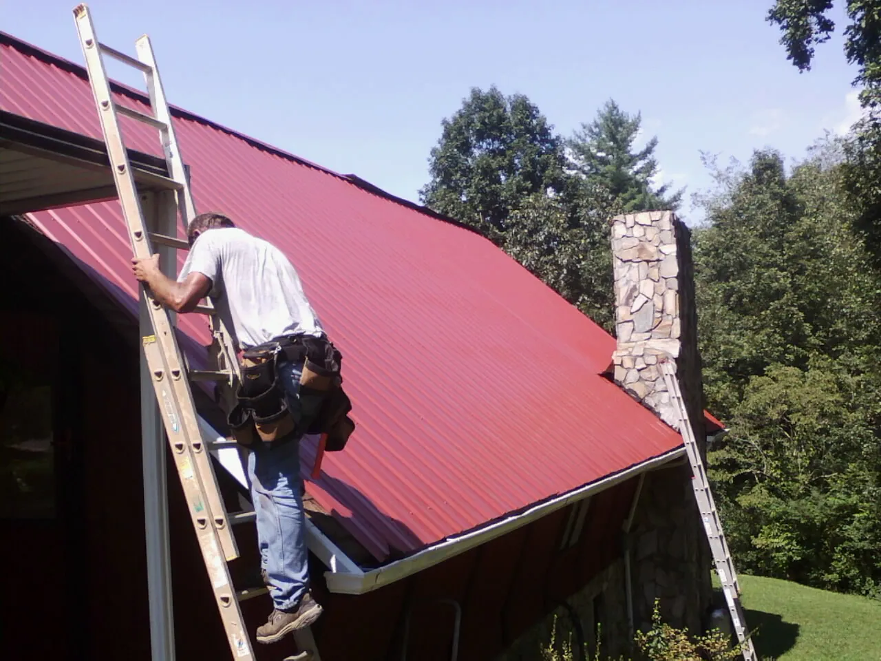 Roofer on a ladder beside a red metal roof and stone chimney — Soffit & Fascia Repair by Roofing Brothers in Pennock
